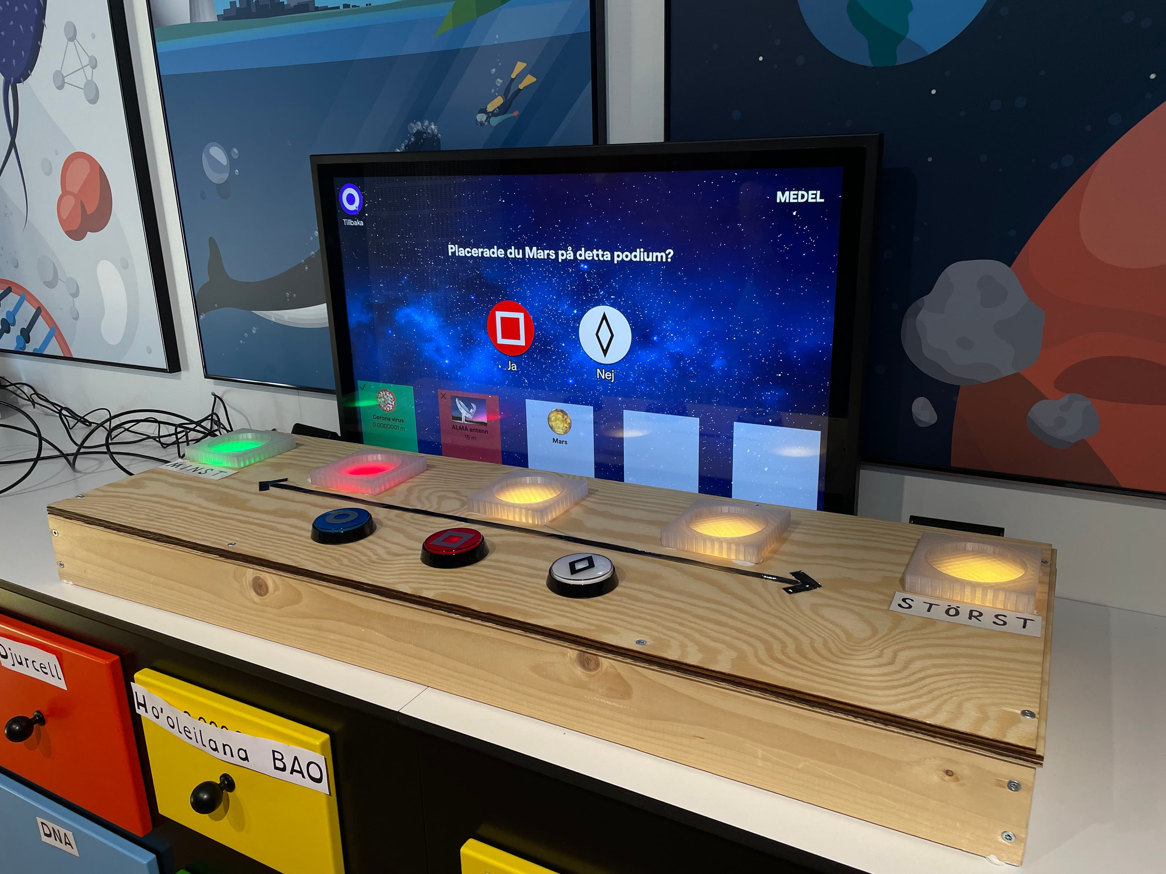 The finished interactive installation at Onsala Space Observatory: a wooden console with buttons and a touchscreen showing a quiz about the scale of scientific objects.